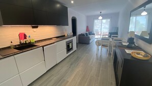 Een moderne keuken met witte kastjes, een zwart aanrechtblad en een inbouwoven. Er is een eethoek met een tafel en stoelen, en een groot raam zorgt voor natuurlijk licht.