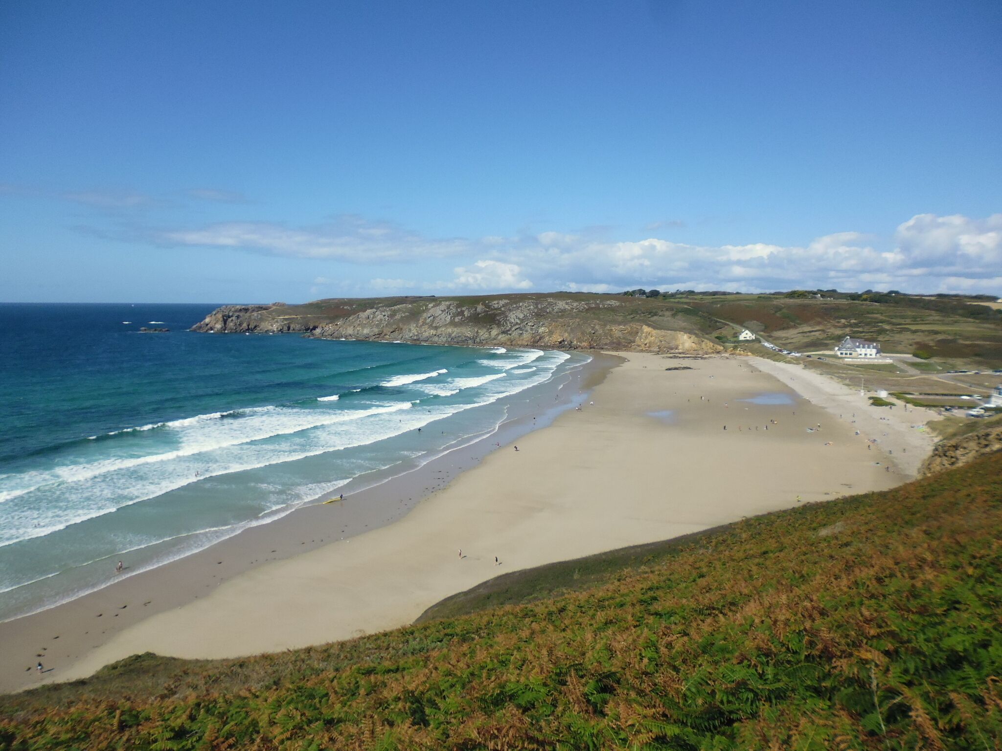 A coastal beach with a sandy shore, a rocky cliff, and a few buildings in the distance.
