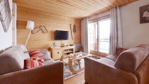 A cozy living room with wooden walls and ceiling, a flat-screen TV mounted on the wall, a wooden coffee table, and two leather sofas with red patterned cushions. - Beautiful apartment/duplex in the Savoyard style in a spectacular setting (Arâches-La-Frasse)