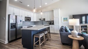 A modern kitchen with white cabinets, stainless steel appliances, a central island with stools, and a dining area with a TV and lamp.