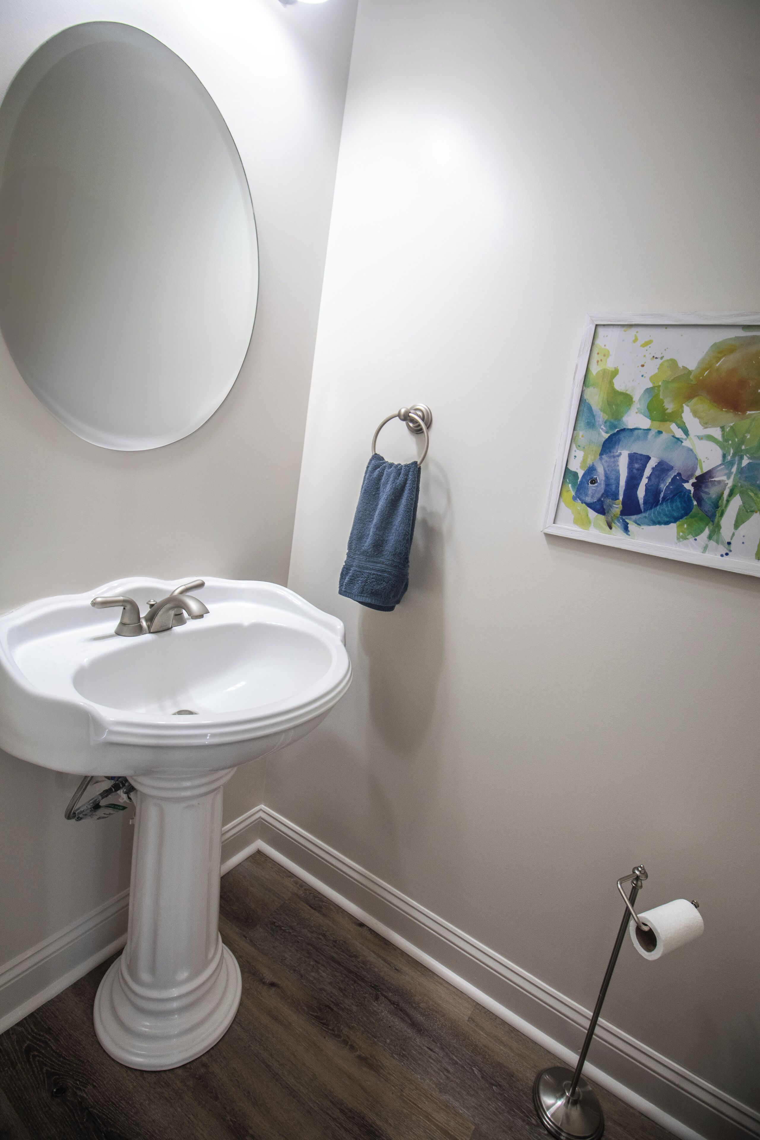 A bathroom with a pedestal sink, a round mirror, a towel rack with a blue towel, a framed artwork, and a toilet paper holder.
