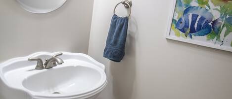 A bathroom with a pedestal sink, a round mirror, a towel rack with a blue towel, a framed artwork, and a toilet paper holder.