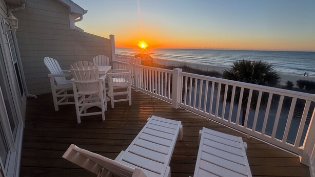A wooden deck with white chairs and lounge chairs, overlooking a beach at sunset.