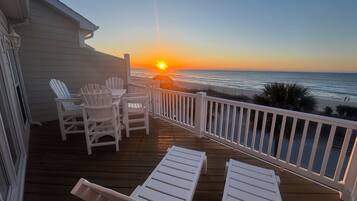 A wooden deck with white chairs and lounge chairs, overlooking a beach at sunset.
