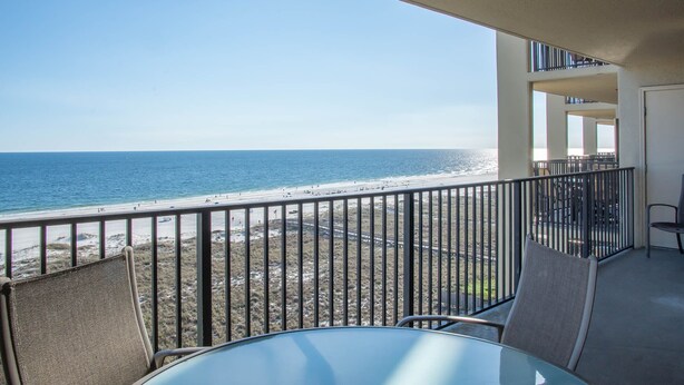 A balcony with a glass table and chairs, overlooking a beach with a clear view of the ocean.