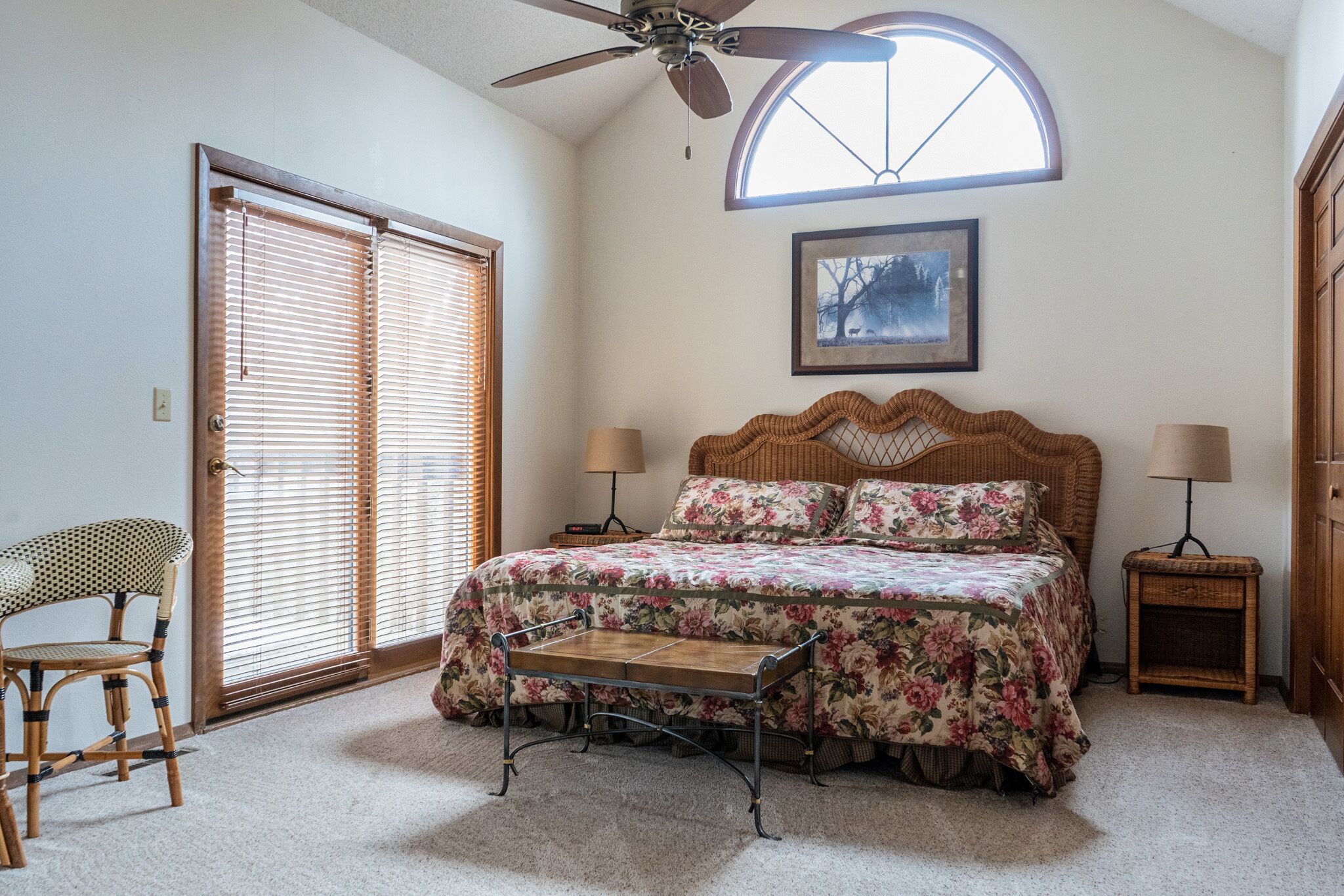 A bedroom with a bed, a chair, a standing lamp, a bedside table, a ceiling fan, and a window with blinds.