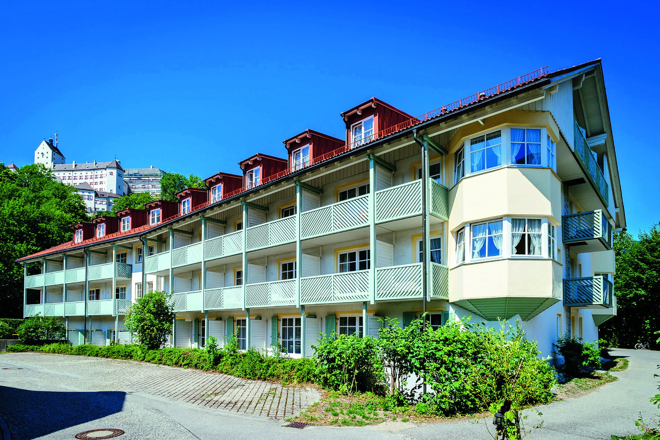 A multi-story hotel with balconies, a red roof, and a view of a cityscape in the background.