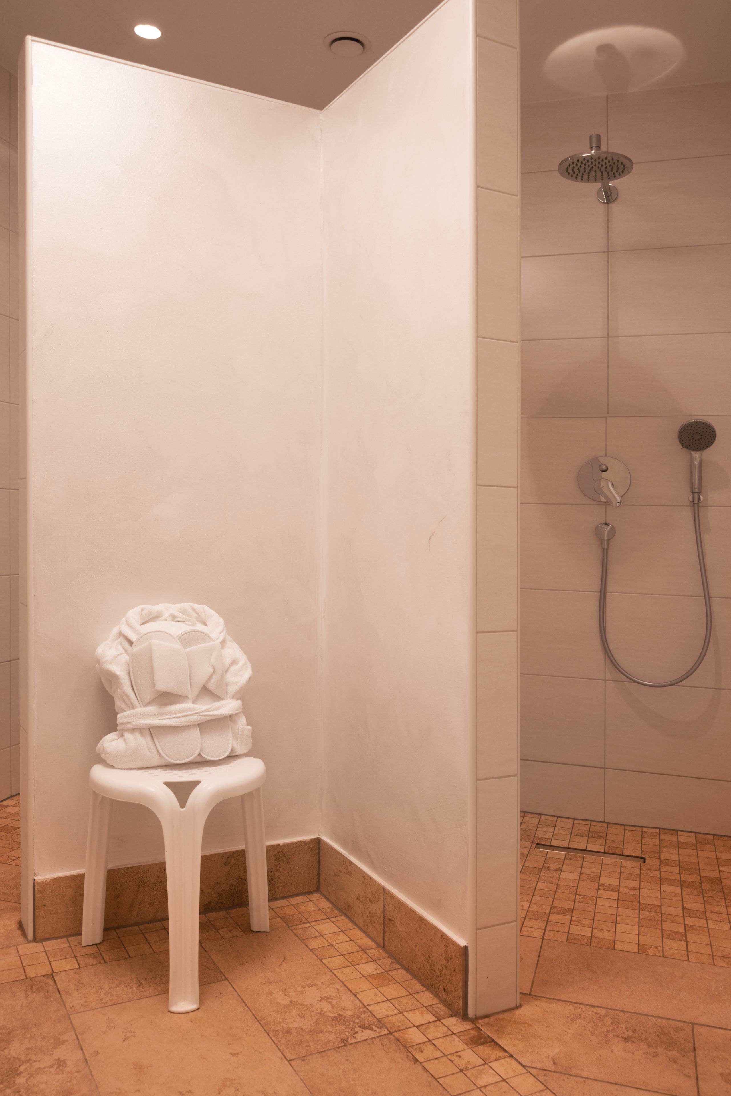 A corner bathroom with a shower, a white chair, and tiled walls.