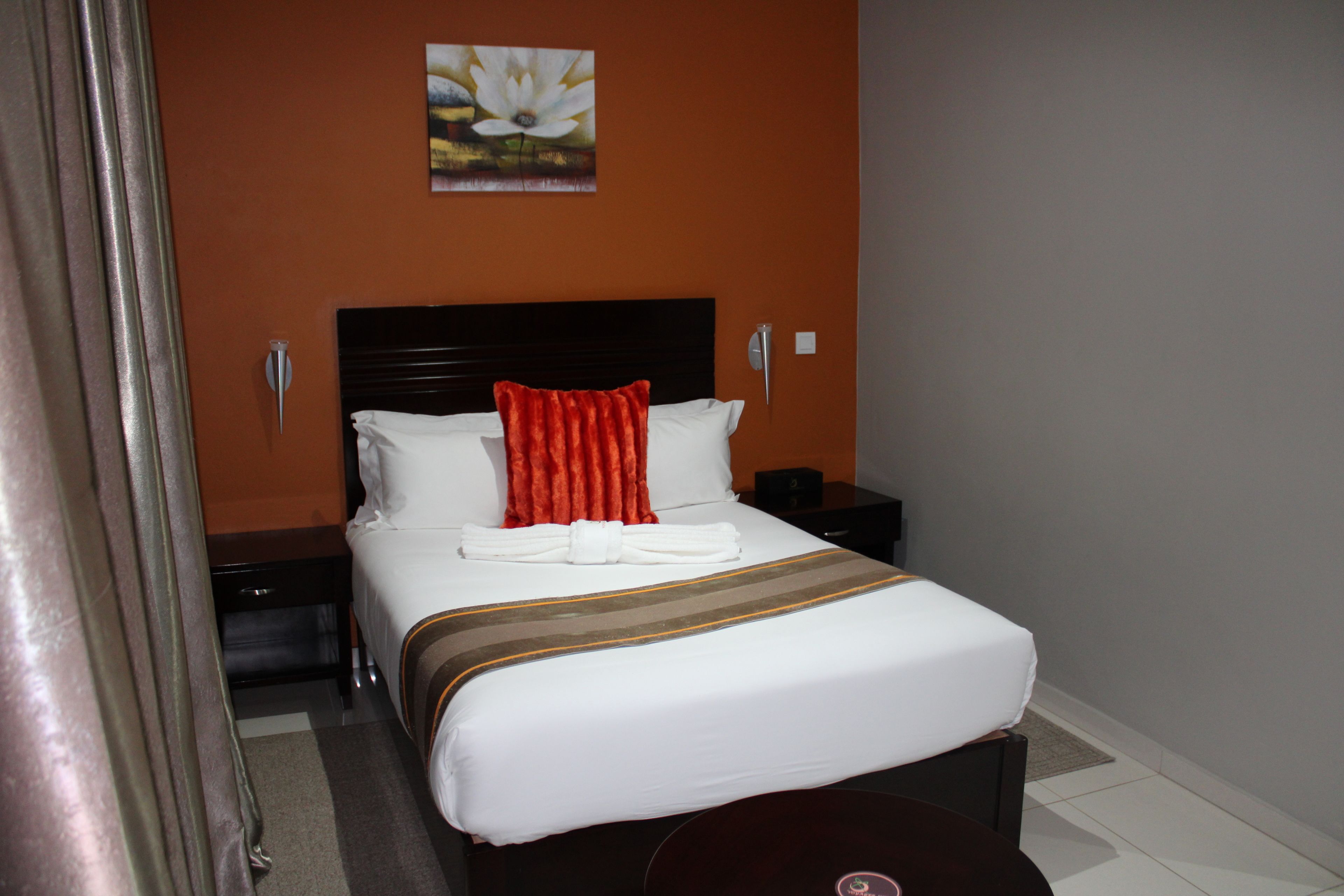 Una cama bien hecha con sábanas blancas y una almohada roja, una mesita de noche con una lámpara y un cuadro de flores en la pared.