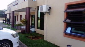 Un bâtiment moderne doté d’une entrée couverte, avec une voiture blanche garée devant et un petit espace aménagé en jardin.