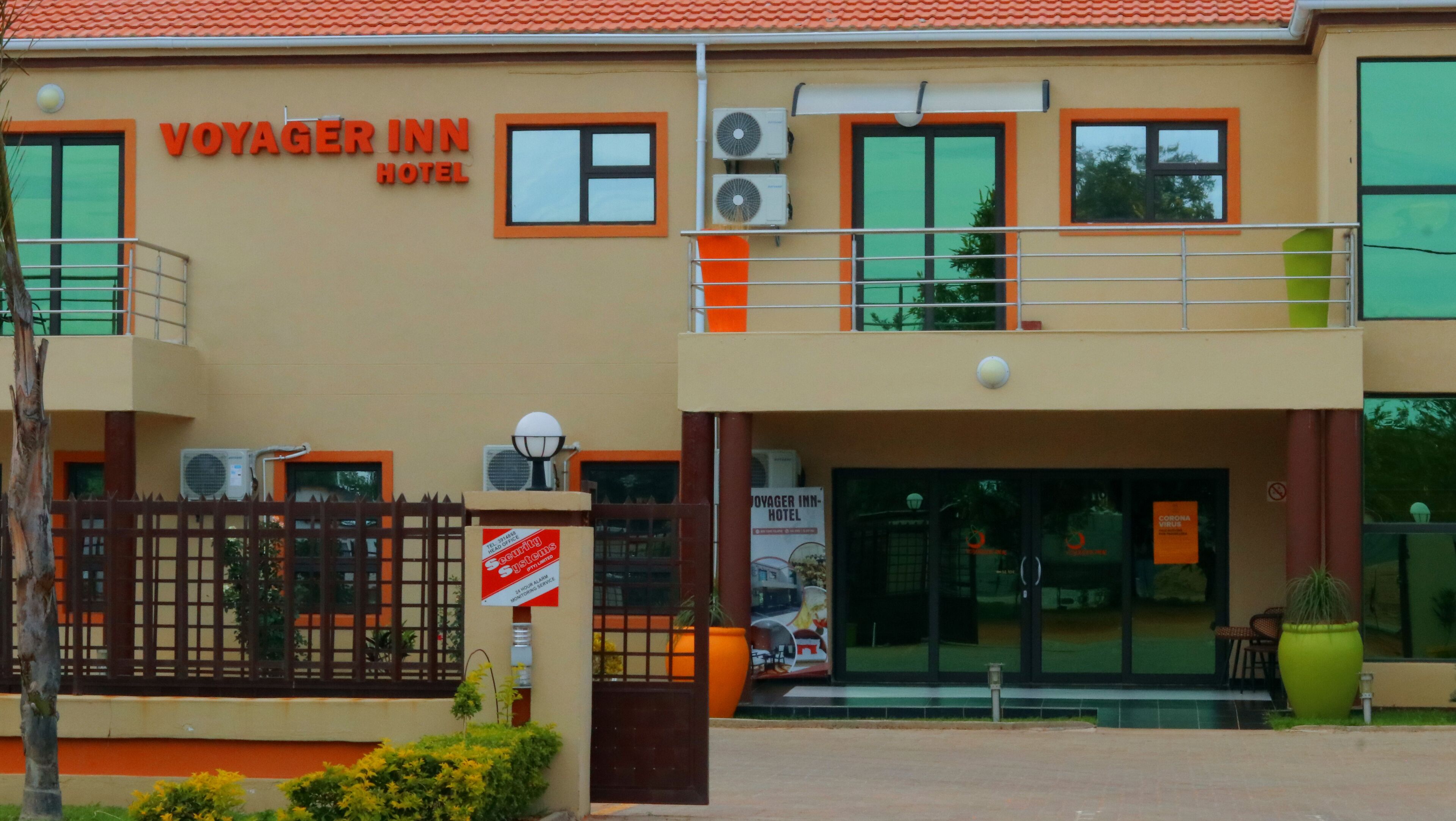 A two-story building with the sign "Voyager Inn Hotel" above the entrance. There are air conditioning units on the exterior walls, a gated entrance, and a small signboard with text.