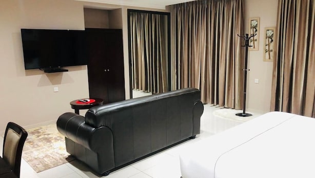 Une chambre d’hôtel avec un grand lit, un canapé noir, un téléviseur à écran plat et une petite table sur laquelle se trouve un objet rouge.