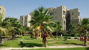 A playground with swings and a slide, surrounded by palm trees and a building with multiple balconies. - The Village Luxury Suites (New Cairo)