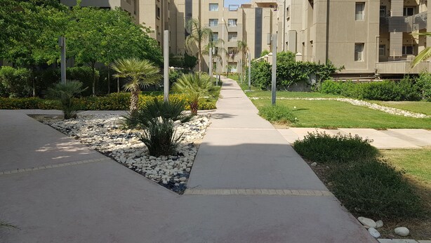 A pathway flanked by modern apartment buildings with balconies, surrounded by greenery and a well-maintained garden area. - The Village Luxury Suites (New Cairo)