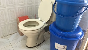 Un inodoro con asiento y tapa color beige, una caja de pañuelos rojos y un recipiente de plástico azul con una etiqueta.