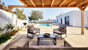 Ein überdachter Außenbereich mit Pool, zwei Korbstühlen, einem Tisch und Blick aufs Meer.