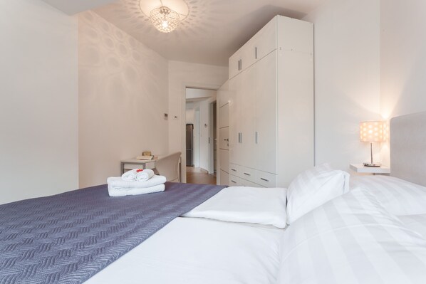 A neatly made bed with white and gray bedding, a small table with a lamp, and a wardrobe with multiple drawers.