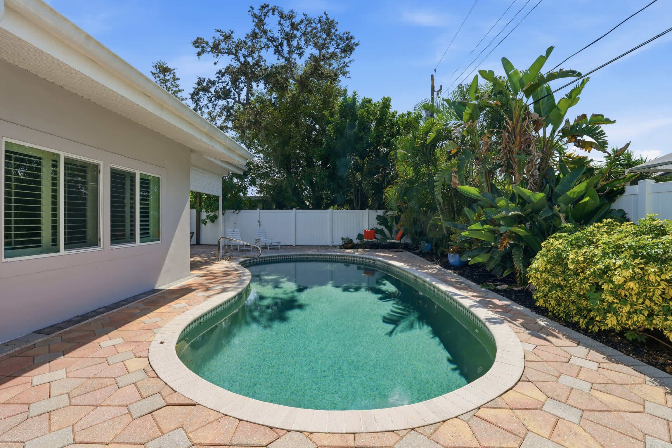 A swimming pool with a tiled deck, surrounded by a white fence, and lush greenery including banana trees and a well-maintained hedge.
