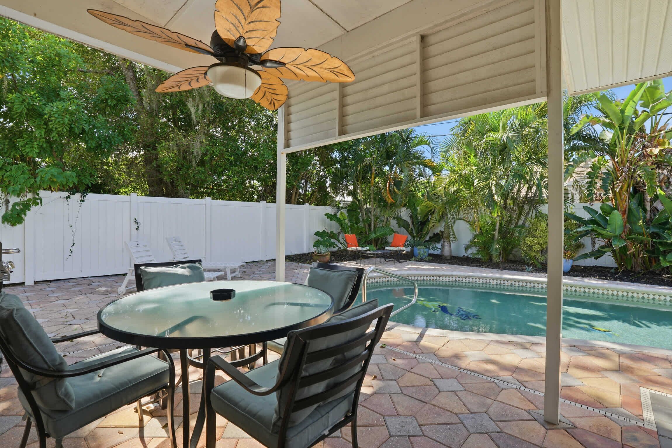 A covered patio with a glass table and chairs, a pool, and surrounding greenery.