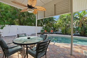 En täckt uteplats med ett glasbord och stolar, en pool och omgivande grönska.