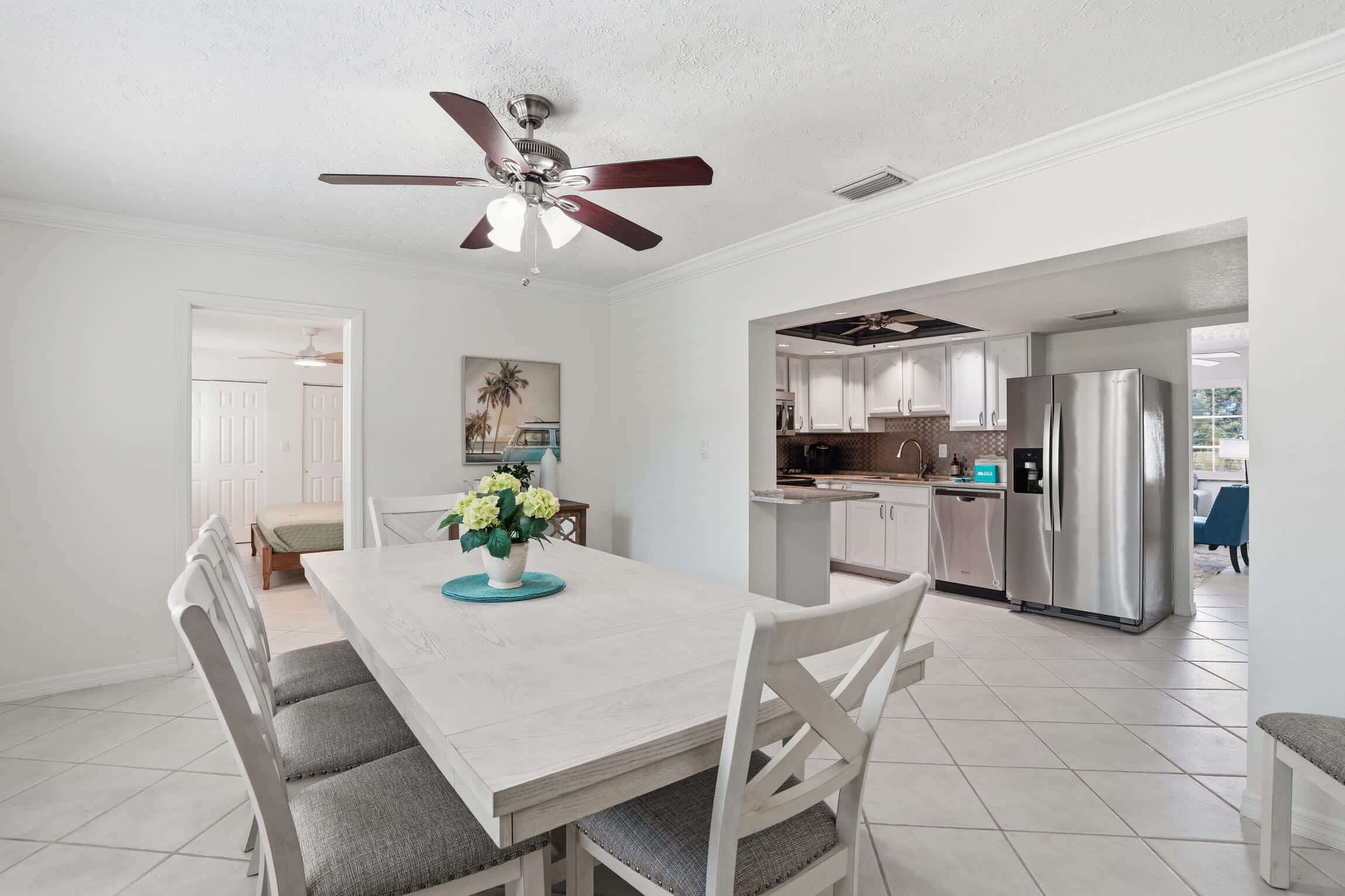 A modern kitchen with stainless steel appliances, a dining area with a white table and grey chairs, and a ceiling fan.