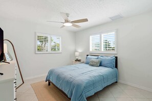 A bedroom with a bed, a ceiling fan, two windows with blinds, a nightstand, and a lamp.