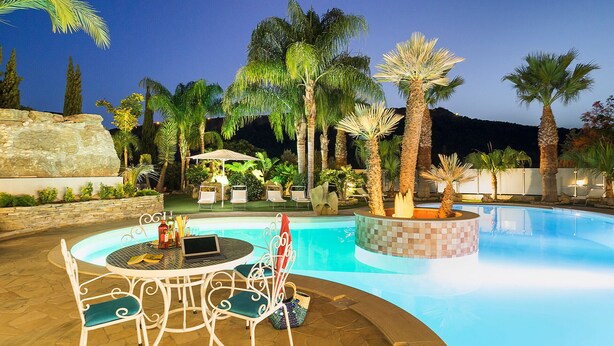 An outdoor swimming pool with a table and chairs, surrounded by palm trees and a stone wall.