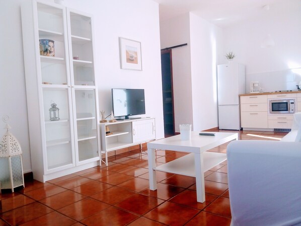 Uma sala de estar moderna com TV, mobiliário branco, uma mesa de jantar e uma área de cozinha com micro-ondas.