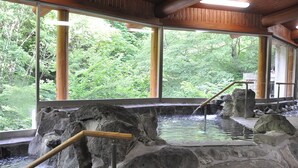 Un onsen japonais traditionnel, avec des bains en pierre, des rampes en bois et une vue sur une végétation luxuriante, visible à travers de grandes fenêtres.