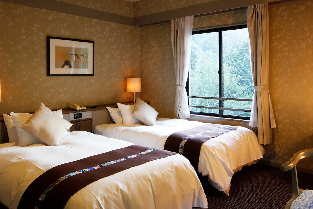Une chambre d’hôtel avec deux lits, un bureau, une chaise, une fenêtre avec des rideaux et un tableau encadré au mur.