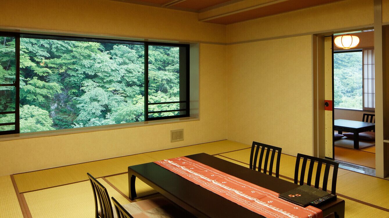 Une pièce de style japonais traditionnel, avec un sol recouvert de tatamis, une table basse agrémentée d’un chemin de table rouge et blanc à motifs, et une grande fenêtre donnant sur un paysage verdoyant et luxuriant.