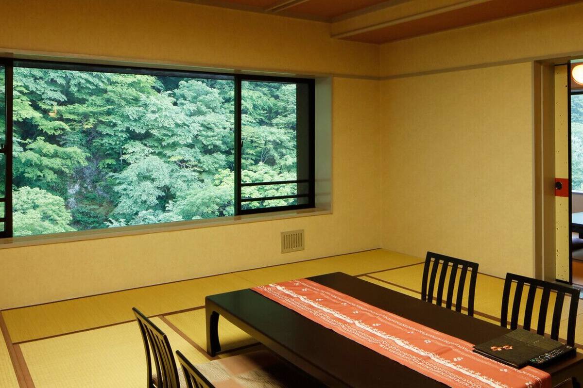 Une pièce de style japonais traditionnel, avec un sol recouvert de tatamis, une table basse agrémentée d’un chemin de table rouge et blanc à motifs, et une grande fenêtre donnant sur un paysage verdoyant et luxuriant.