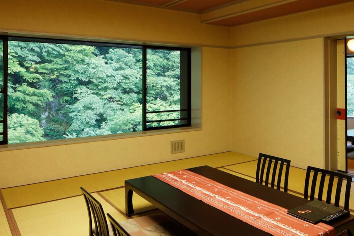 Une pièce de style japonais traditionnel, avec un sol recouvert de tatamis, une table basse agrémentée d’un chemin de table rouge et blanc à motifs, et une grande fenêtre donnant sur un paysage verdoyant et luxuriant.