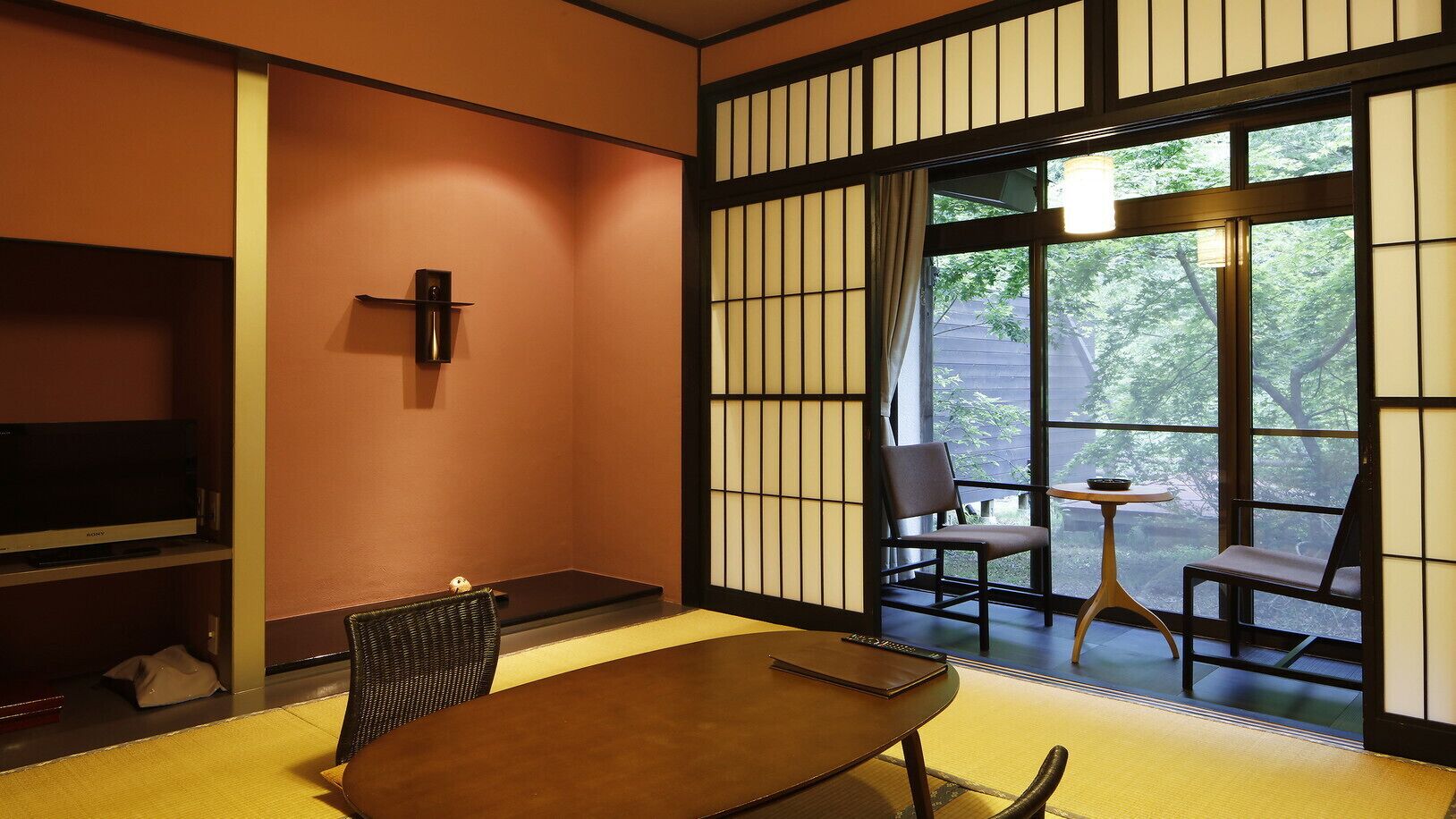 Une pièce de style japonais traditionnel, avec un sol recouvert de tatamis, une table basse, des chaises, un téléviseur et une porte coulissante donnant sur un paysage verdoyant.