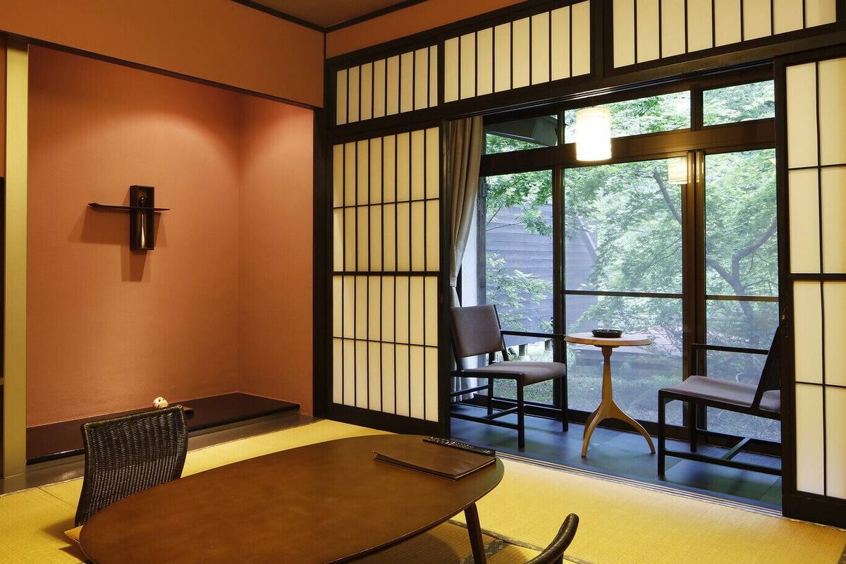 Une pièce de style japonais traditionnel, avec un sol recouvert de tatamis, une table basse, des chaises, un téléviseur et une porte coulissante donnant sur un paysage verdoyant.