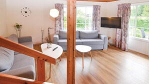 A modern living room with a wooden staircase, grey sofas, a flat-screen TV, and large windows with floral curtains. - 2 bedroom pet friendly home in Broager (Broager)