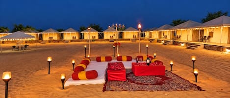 A desert-themed outdoor seating area with cushions and tables, surrounded by tents and illuminated by candles.