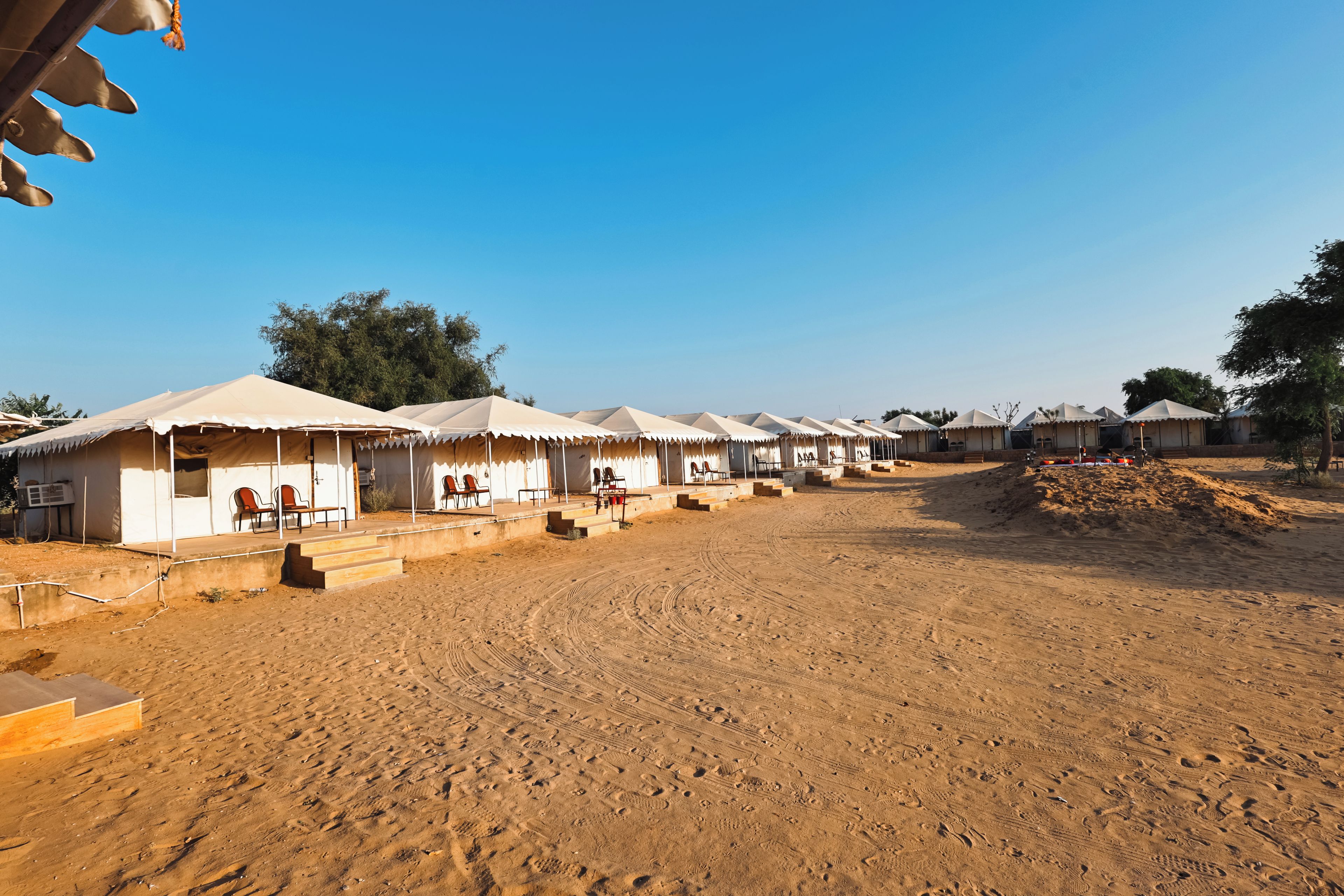 A row of tents with steps leading to the entrance, set in a sandy area with trees in the background.