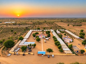 A large, open-air facility with multiple tents and a central colorful structure, surrounded by sparse vegetation and a flat landscape. - Rajputana Desert Camp By Castle Crown (Jaisalmer)