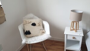 Ein weißer Stuhl mit einem flauschigen Sitz und einer beige farbenen Decke mit Fransen, ein weißer Nachttisch mit einer Lampe und ein Bett mit Punkten.