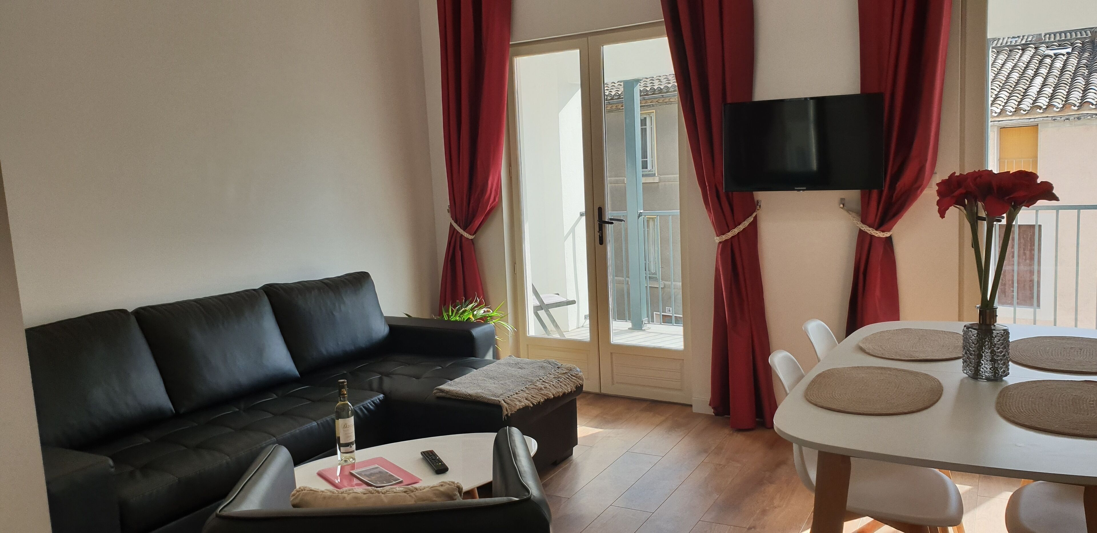Ein Wohnzimmer mit einer schwarzen Couch, einem Couchtisch mit einer Flasche und Fernbedienung, einem Esstisch mit Platzsetzern und einem Blumenstrauß sowie einem an der Wand montierten Fernseher.