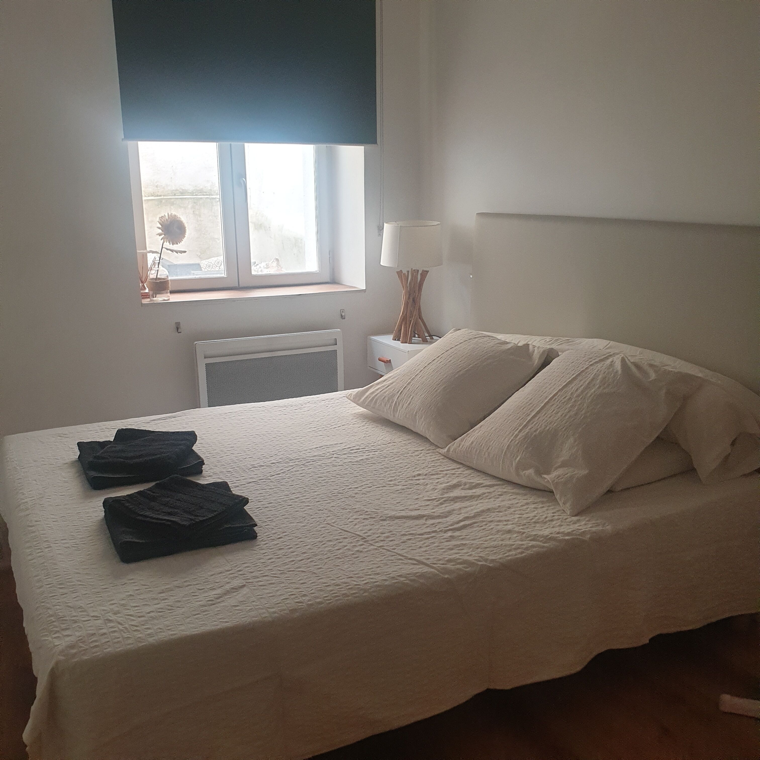 Ein ordentlich bezogenes Bett mit weißer Decke und Kissen, eine Nachttischlampe und ein Fenster mit Blick nach außen.