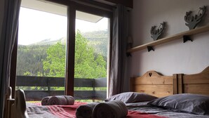 A bedroom with a wooden headboard, a bed with rolled towels, a red and white patterned bedspread, and a view of a forest through a large window. - Apartment 4/6 pers, 50m², Les Houches Chamonix (Les Houches)