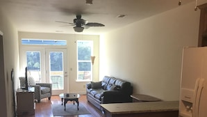 A spacious living area with hardwood flooring, a black leather couch, a small coffee table, and a white refrigerator.