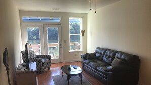 A living room with a black leather couch, patterned armchair, and a small table. There is a sliding glass door leading to an outdoor patio.