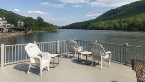 A deck with wicker chairs and a table overlooking a calm river, surrounded by lush greenery and hills.