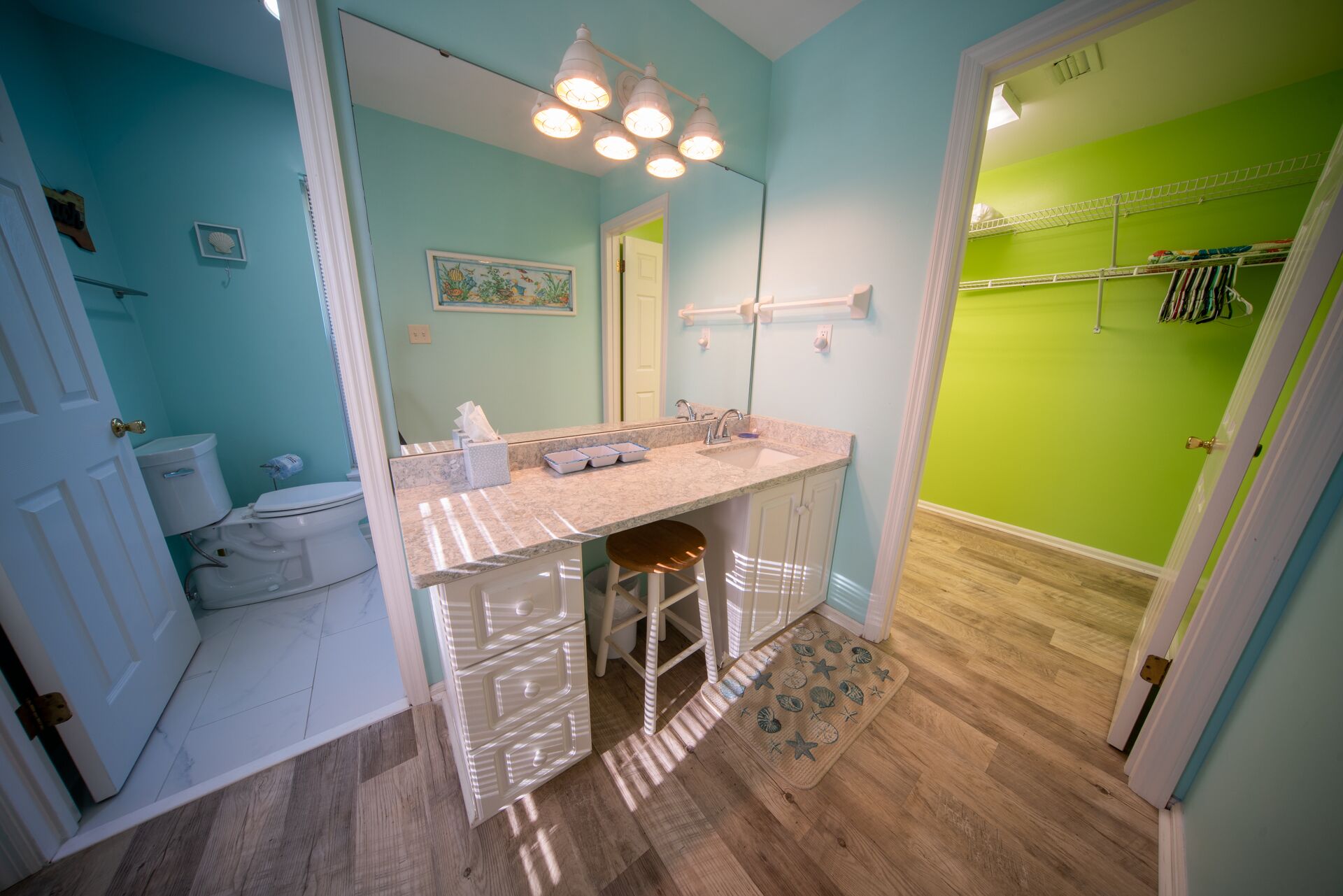 A bathroom with a vanity, sink, and toilet. There is a mirror above the sink and a towel rack above the toilet. The walls are painted in different colors, and there is a wooden floor.