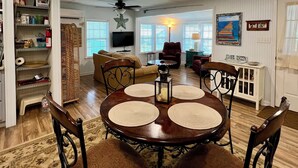 A dining area with a round table, four chairs, and a chandelier. There is a television in the background, and a 'WELCOME' sign on the wall.