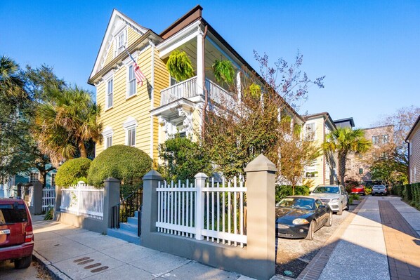 A two-story house with a balcony, a white picket fence, and a driveway with parked cars.