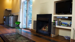 A living room with a fireplace, a television mounted on the wall, and a kitchen area with a refrigerator and a kettle. - Marwood Villas (Halls Gap)